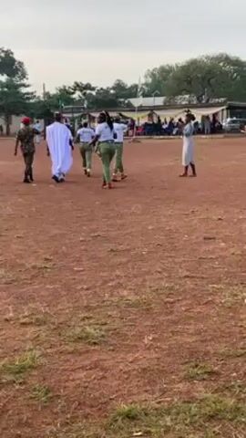 The video shows a group of people walking across a dirt field. One man is wearing a white robe, another is in military uniform, and several women are wearing green and white outfits. The group is walking toward a crowd of people sitting under a tent. There are some trees and buildings in the background, and the sky is cloudy.
