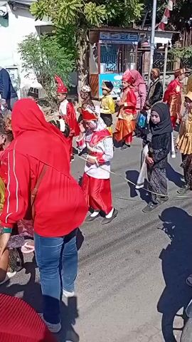 A procession is taking place on a street. A group of children, dressed in traditional attire, are marching. Some are wearing red and gold costumes, while others are in yellow and black. One child is wearing a black hijab and a patterned skirt. A woman in a red hijab and blue jeans is standing on the left side of the frame, facing away from the camera. Another person, possibly a child, is sitting on a motorcycle in the foreground, wearing a pink hoodie and looking towards the procession. A red motorcycle helmet is visible on the back of the motorcycle. Trees and buildings line the street, and a flag is visible on a pole in the background. The overall atmosphere appears festive.
