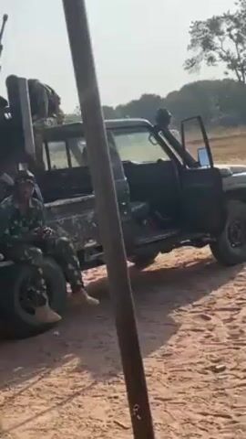 The video shows several armed soldiers in a sandy outdoor environment. Some are sitting in the back of a truck fitted with a machine gun, while others are walking around. The soldiers are wearing camouflage uniforms. In the background, there are trees and a building. The weather appears to be sunny.