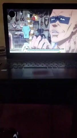 The video shows a laptop screen displaying a scene from an anime, showing a man with blue face paint holding something in his hands, and another man with long blue hair. The laptop is sitting on a dark surface.