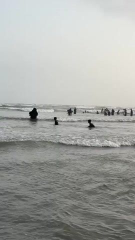 This media file captures a scene at a beach, likely in the Karachi Division of Pakistan, given the provided location context. The main subjects are a group of people enjoying the water. Several individuals are partially submerged, indicating they are swimming or wading. In the foreground, the waves are breaking, creating white foam and demonstrating the motion of the sea.

The time of day appears to be overcast, possibly late afternoon or early morning, given the muted light and the absence of harsh shadows. The weather seems to be cloudy, contributing to the diffused lighting. The overall mood of the scene is relaxed and recreational, with people engaging in typical beach activities.

In the mid-ground, more people are visible further out in the water. One individual in the left mid-ground is wearing what appears to be a dark abaya or religious attire, standing in the water. There are also several figures scattered further back, suggesting a moderately populated beach. The background is dominated by the ocean meeting the sky, with no distinct landmarks visible to pinpoint the exact location.

The scene is devoid of any visible text. The focus is solely on the natural environment of the beach and the people interacting with it. The gentle rolling of the waves and the presence of people in the water suggest a tranquil, yet engaging, atmosphere.