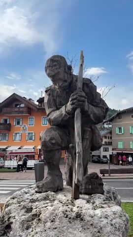The video shows a statue of a man carved from wood, kneeling and holding a wooden staff. The statue is on a large rock. In the background are buildings with balconies and flowers, and people walking on the street and sitting on benches. The sky is blue with some clouds. There are also some temporary barriers on the side of the road. The scene appears to be in a town square.