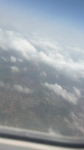 The video shows an aerial view from an airplane window. Below, a city is partially obscured by scattered clouds. Above, a vast expanse of white clouds extends towards a light blue sky. The edge of the airplane window frame is visible at the bottom.