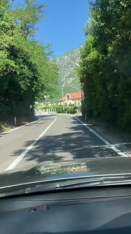 A camera mounted on a car dashboard records a drive along a paved road. The road curves gently to the left, marked with a white solid line in the center. Trees line both sides of the road, creating a tunnel-like effect in some sections. A building with a red tile roof and a restaurant sign sits off to the right side of the road. Beyond the building and trees, a mountain looms in the background, covered in green vegetation and patches of exposed rock. The sky is clear and blue. As the car continues, a car parked by the side of the road can be seen and Croatian flags can be seen flapping in the wind.