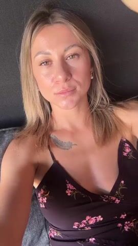 A woman with blonde hair and a floral dress is lying on a dark gray couch, looking at the camera. Her left hand is touching her hair and her right hand is touching her face.