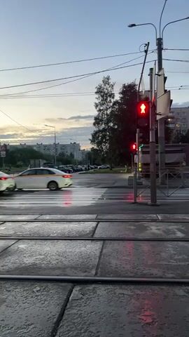 This video captures a street scene at dusk in Ladozhskoe Ozero, Russia. The sky is a dusky blue with hints of orange and pink from the setting sun, suggesting it is either early morning or evening. The weather appears to be damp, as evidenced by the wet road surface and reflections on the asphalt.

The focal point of the video is a pedestrian crossing with a traffic light. The pedestrian signal displays a red "don't walk" symbol, indicating that it is not safe for pedestrians to cross. The vehicular traffic light is also red. Several cars are visible on the road, with one white car and a dark-colored car in motion, their headlights creating streaks of light due to the slow shutter speed. In the background, apartment buildings and trees line the horizon. A parked line of cars can be seen to the left of the frame. The presence of tram tracks in the foreground suggests that this is a streetcar route. The overall mood is calm and a bit melancholic, typical of a quiet evening in a city. There is no visible text in the video.