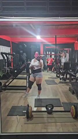 The video shows the reflection in a gym mirror of a person taking a selfie. The person is standing to the left of a barbell, wearing a white top, grey shorts, a waist belt, knee pads, and sneakers. They are holding their phone in their left hand, presumably to take a picture of themselves. In the background of the gym, other people are working out on various weight machines. The gym has wood-look flooring, and the walls are primarily black and red. The lighting is bright.