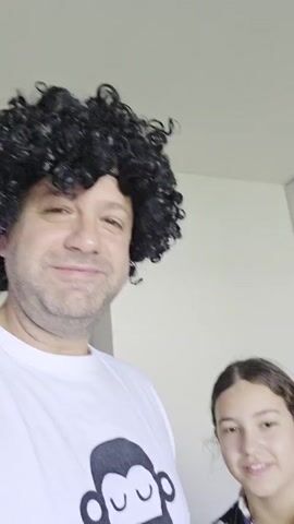 A man wearing a curly black wig stands next to a young girl with long brown hair. Both are smiling. The man is wearing a white t-shirt with a black graphic. They are indoors in front of a white wall with a white cabinet. A small round wall decoration hangs on the wall.