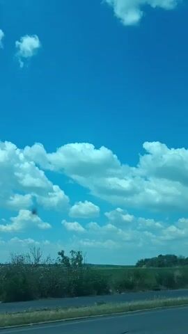 The video shows a view from a vehicle moving along a road. The sky is bright blue with scattered white cumulus clouds. In the distance, there is a flat, green landscape, likely fields, with a line of trees on the horizon. The foreground shows a road surface with a strip of dry grass and brush along the side. There is no visible indication of people or specific actions taking place. The overall impression is of a scenic drive through a rural area on a clear day.