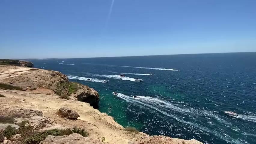 A high-angle, wide shot captures a vast, blue ocean under a clear, bright sky. The foreground is dominated by a rugged, rocky cliff face, bathed in sunlight, with sparse green vegetation. The sea below the cliff is a vibrant turquoise, transitioning to a deep blue further out. A fleet of approximately ten white speedboats is scattered across the water, each leaving a white wake as they move in various directions, suggesting a recreational event or a guided tour. Some boats are closer to the cliff, while others are further offshore. The distant horizon shows a hazy coastline or landmass. The overall impression is a sunny day at sea with boating activity.