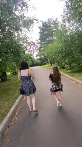 Two young women are walking away from the camera on a paved path in a park. They are surrounded by green trees and grass. In the background, there is a large Ferris wheel with red supports. The woman on the left is wearing a black and white polka dot dress and carrying a black purse. The woman on the right is wearing a plaid shirt over a black shirt and shorts, and white sneakers. She is holding a pink balloon and has her arms raised, as if throwing something. The Ferris wheel appears to be old and possibly disused. The sky is overcast.