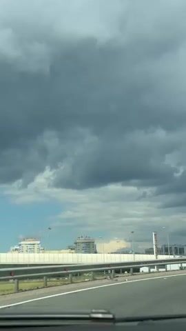 The video shows a view from inside a vehicle driving on a highway. The sky is filled with dramatic, dark storm clouds, contrasted with patches of blue sky and white clouds. In the distance, there are modern buildings, tall streetlights, and a highway barrier. A red and white directional sign indicating a sharp curve to the right is visible on a pole. The road surface is asphalt with white lane markings. The windshield wipers are visible at the bottom of the frame. The overall impression is of driving towards or away from a storm.