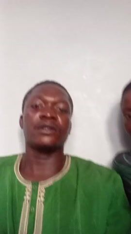 The video features two individuals against a white background. The primary focus is on a man wearing a green shirt with embroidered detailing around the neckline. He is facing the camera, speaking, and gesturing with his right hand. Beside him, slightly behind and to the right, is another person wearing a dark head covering and striped shirt. Their face is partially visible. The environment is simple, focusing attention on the two people.
