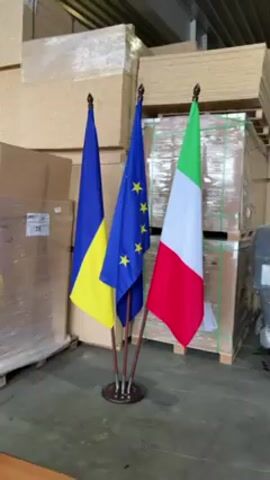 The video shows a floor stand displaying the national flags of Ukraine, the European Union, and Italy. The Ukrainian flag is on the left, with blue over yellow. The EU flag is in the middle, with a blue field and yellow stars. The Italian flag is on the right, with green, white, and red stripes. The flags are set against a backdrop of stacks of packaged wooden boards. The floor is concrete. There are boxes and other materials in the background. The light is artificial. The camera is stationary.