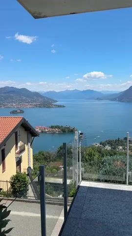 A vertical video shows a scenic view of a large body of water surrounded by hills and mountains under a bright blue sky with scattered white clouds. In the foreground, a section of a modern building with glass railings and a tiled patio is visible. To the left, a yellow house with a red-tiled roof sits on a slope, bordered by a black fence. The middle ground features a picturesque scene with several islands in the water, some with buildings and lush greenery. Small boats are scattered across the water. The background reveals rolling hills and distant mountains that fade into the horizon. The overall environment appears to be a resort or residential area in a Mediterranean-like climate. There are no people visible in the video.