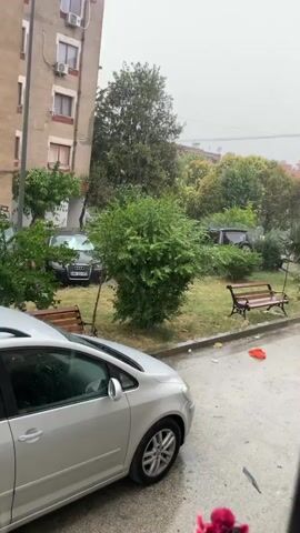 A video shows a street scene during a rainstorm. A silver car is parked on the left side of the frame, facing away from the camera. In the background, there are trees, a building with windows, and two park benches. It appears to be daytime, but the sky is overcast. The rain is visible hitting the ground and the cars.