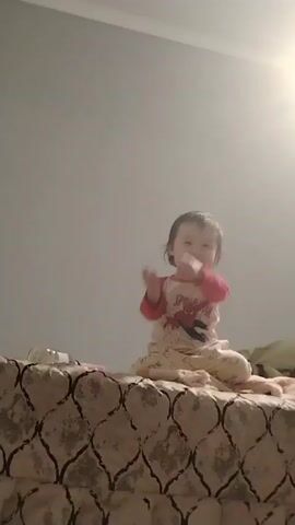A toddler sits cross-legged on a bed with a brown and beige patterned cover. The child wears a red and white long-sleeved shirt with a Spider-Man design and light-colored pajama pants. The toddler waves a hand, speaks, and seems to be looking at someone or something off-camera. The room is brightly lit and the background is a plain gray wall. Some additional items such as a pillow and what appears to be a humidifier are on the bed.