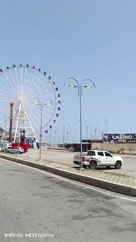 The video shows an outdoor amusement park scene with a large Ferris wheel as the central subject. The Ferris wheel is white with colorful gondolas. In the foreground, there's a road with several cars and trucks parked along the side. A red tent is also visible. The setting appears to be a sunny day with a clear blue sky. The Ferris wheel is not in motion. There are streetlights and some fencing in the area. The overall impression is a lively, possibly coastal, amusement park. The bottom of the screen displays "Infinix HOT 50 Pro", indicating the device used to record the video. There are no discernible people in the video, nor any specific event taking place, other than the general presence of an amusement park.