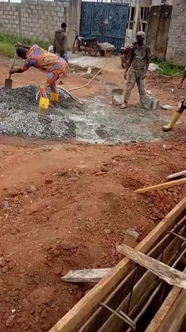 In an outdoor construction setting, a person wearing a colorful patterned shirt, orange pants, and yellow boots is actively mixing a pile of gravel and cement with a shovel. A hose is visible near the pile, suggesting water is being used in the mixing process. In the background, several other individuals are present. One person, wearing a white shirt and dark pants, stands near the mixing area holding a white object. Further back, another person in a dark shirt and blue shorts is near a wheelbarrow. To the left, two people are walking away from the camera. The ground is composed of reddish-brown soil with scattered rocks and debris. A fence and part of a building are visible in the distance, as well as a motorcycle.