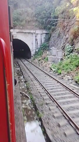 The video shows a train moving along a track, passing through a tunnel. The view is from inside the train, looking out the side. The train is red, and the tracks are steel. The tunnel is dark, and the walls are lined with stone. As the train exits the tunnel, lights can be seen lining the walls. The surrounding environment is dark.