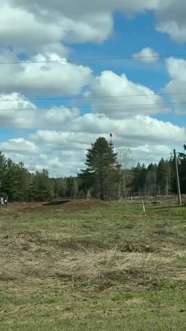 The video presents a daytime view from a moving vehicle, capturing a rural landscape in Сундур, Russia. The sky is bright blue with scattered, fluffy white clouds.

In the initial frames, a large grey utility pole occupies the right side of the foreground, with power lines extending across the top of the frame. Beyond the pole, a field of dry, brownish grass with patches of green stretches into the midground, leading to a line of sparse trees, including both evergreens and deciduous trees, some of which are bare.

As the camera pans, a tall, dark metal flare stack becomes visible in the distance, situated amidst the trees. A large, vibrant orange flame burns actively at the top of the stack, indicating gas flaring. To the left of the flare stack, a piece of yellow industrial equipment is partially visible, and a small white sign with illegible text briefly appears. The scene suggests an industrial operation, likely related to oil or gas production, set within a natural, wooded environment. No people are visible in the video. The time of day is clearly daytime with bright, sunny weather.