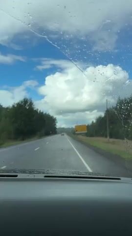 The video shows a view through a car windshield. The windshield has raindrops on it, blurring the view slightly. The road ahead is wet and appears to be a highway with two lanes in each direction. There are trees lining both sides of the road, and a yellow sign is visible on the right side. The sky is blue with large white clouds. There are no people or other vehicles clearly visible in the shot. The car appears to be in motion.
