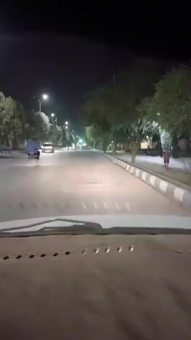 A first-person perspective video shows a car driving down a street at night. Streetlights illuminate the road and trees lining the sidewalk. There are a few vehicles and people visible on the road and sidewalk. A motorbike is visible ahead on the road. The car is traveling on the right side of the road, and there is a sidewalk on the right with some benches and a trash can. The video captures the car's dashboard in the foreground.