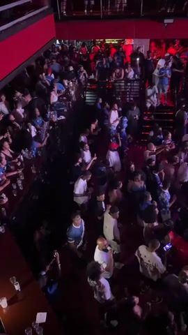 A dimly lit concert venue is filled with a large crowd of people standing and dancing. The crowd is dense, with individuals of various ethnicities and ages. Many are dressed casually. In the background, a stage is visible with a DJ setup and a large screen displaying an American flag with stars and stripes. Lighting is predominantly red and purple, with some spotlights. Several rows of seating or balconies overlook the main floor. The atmosphere appears lively and energetic.