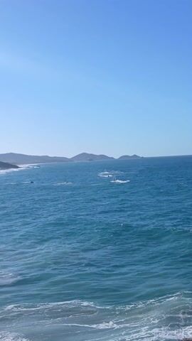 The video shows a scenic view of the ocean and a beach. Initially, the focus is on the ocean with several boats visible in the distance. As the camera pans right, it reveals a sandy beach with people swimming and relaxing along the shoreline. In the background, there are mountains covered with trees, possibly palm trees, and a few buildings are visible. The sky is blue with some scattered clouds, and the sun is shining brightly, causing a slight glare on the water. The waves are crashing on the beach, creating a white foamy edge.