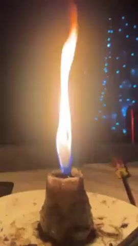The video shows a burning candle on a white plate against a dark background, with blue Christmas lights visible in the distance. The flame is tall and narrow, flickering in the dark. The candle itself appears to be thick and possibly homemade, with a rough, uneven surface. The plate is slightly dirty, with wax drippings and other debris visible. A tool is laying to the right side of the plate, only partly visible. There are no people visible in the video. The scene is shot in low light, creating a warm and cozy atmosphere.
