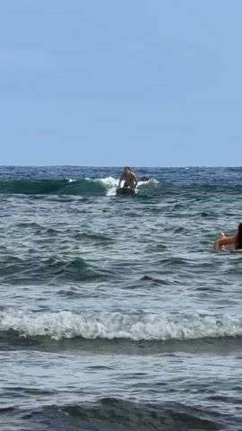 The scene shows a person surfing in the ocean on a bright, clear day. The individual, dressed in swimwear, starts crouched on the surfboard, then stands up before losing balance and falling into the water. The ocean has moderate waves and the sky is a clear blue. Another person's arm is partially visible in the foreground.
