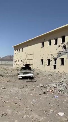 The video shows a two-story tan-colored building riddled with bullet holes and significant structural damage. Windows are missing and large chunks of the building's facade are destroyed. Debris litters the ground around the building, including pieces of concrete and metal. A completely destroyed car sits in front of the building, its charred remains visible. The sky is clear and blue, suggesting a daytime setting. Sandbags are visible in front of the entrance to the building.