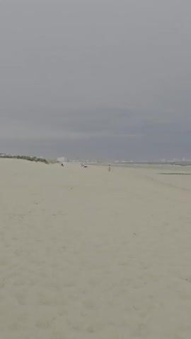 The video shows a wide, sandy beach under a cloudy sky. In the distance, there are small figures of people and possibly some structures or buildings along the coastline. The sea is visible as a calm, gray expanse meeting the horizon. The sand appears mostly flat with some tire tracks visible in places. There are no discernible objects or significant actions happening in the foreground.