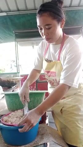 A woman with her hair tied up in a bun, wearing a white t-shirt and a yellow apron with red trim, is standing in what appears to be an outdoor food stall. She is wearing white disposable gloves and is using a pair of metal tongs to transfer food from a green bucket to a pink colander that is sitting inside a blue bowl. The food appears to be small, irregularly shaped pieces of a light-colored mixture, possibly dough or batter. There are other food preparation items visible in the background, including another green bucket, a red cabinet, and what looks like a stove or cooking apparatus. The lighting suggests it is daytime.