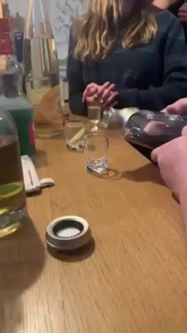 The video shows a person pouring a dark amber liquid from a bottle labeled "De Luze Cognac" into a small shot glass on a wooden table. There are three other shot glasses on the table, two containing clear liquid and one appearing empty. Other bottles, including one labeled "Génépi", are visible in the background. Another person sits across the table, hands folded in front of them, wearing a green and blue plaid sweater. A silver bottle cap sits on the table.