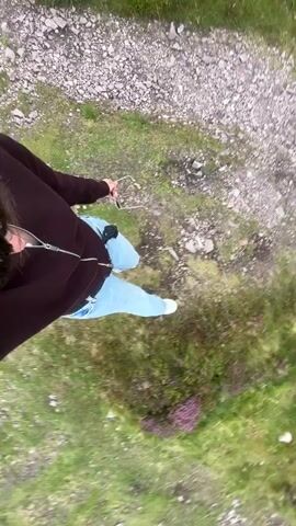 This video shows a person's legs and feet from a high angle, looking down. The person is wearing light blue pants and a dark brown hoodie. They are standing on a rough path that is made of small rocks and gravel. To the right of the path, there is green grass and some purple flowering plants. The person is walking on the path, and their right leg is extended forward. The camera appears to be attached to the person's head or upper body.