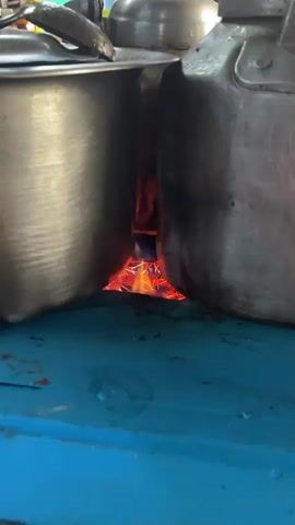 The video shows a close-up of two metal pots placed side-by-side over an open flame. The flame, visible between the pots, glows with intense orange and red hues, indicating high heat. The pots are positioned such that their lower halves are exposed to the direct heat source, suggesting they are being used for cooking or heating something. The surrounding surface appears to be a blue, possibly wooden or painted material, with some water droplets visible. The scene is dark and focused on the fire and the pots.
