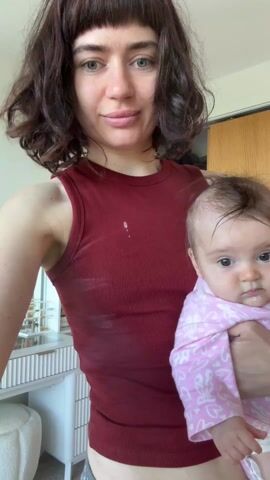 A woman with brown hair, wearing a red tank top and plaid shorts, is holding a baby wrapped in a pink blanket. The baby is looking at the camera. The woman smiles slightly at the camera, then rotates slightly to her right. Behind her is a white vanity table with makeup, a closet with open doors, and a white plush rabbit.
