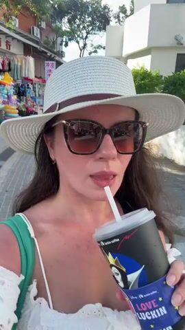 A woman with long dark hair, wearing a white hat and sunglasses, is holding a black and purple cup with a straw and drinking from it. She is wearing a white eyelet top with green straps and a brown purse is visible. The background shows a street with shops on the left and trees and a white building on the right. The ground is paved. The woman is looking towards the camera.