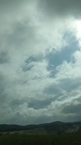 The video shows a time-lapse of a cloudy sky over a rural landscape. The sun is visible in the upper right corner, peeking through the clouds. The clouds appear to be moving, with some patches of blue sky visible between them. The landscape below consists of rolling hills covered in trees and fields. The overall impression is one of a dynamic and changing sky.