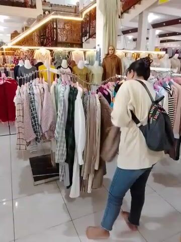 The video shows a woman in a store, likely a clothing store, browsing through racks of clothes. She is wearing jeans and a light-colored shirt, and has a backpack on. She is barefoot. In front of her are several racks of clothing, mostly shirts and blouses. Behind her, there are more racks of clothes and mannequins displaying clothing. The store appears to be well-lit and spacious, with tiled floors and a decorative ceiling. The woman is examining a beige shirt on a hanger.