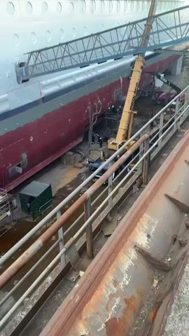 The video shows a shipbuilding or ship repair scene. A large section of a ship hull, painted red with white accents, is visible in a dry dock. A yellow MEDIAGCO crane is centrally located, with its boom extended towards the ship's hull. A person is visible in a work platform attached to the crane, welding the ship. Sparks are visible from the welding. Several people, wearing hard hats and safety gear, are working around the base of the crane and the ship. Scaffolding is also set up along the hull. In the foreground, a rusty metal railing provides a vantage point for the camera. A small green portable building is visible on the ground near the hull. Another crane, labeled "M REN," is partly visible in the background. A pink utility vehicle sits nearby. The setting suggests a busy industrial environment focused on the construction or maintenance of large vessels.
