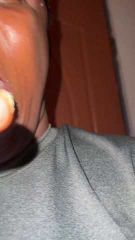 The video shows a person with dark skin, wearing a gray shirt. They hold a piece of food up to their mouth and take a bite. The background shows a brown door, a tile floor, and an old-fashioned sewing machine. The floor also has a pair of flip-flops on it. The lighting appears to be indoor, and the camera angle is close-up and slightly angled.
