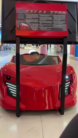 A young boy with short, dark hair and a light-colored hat is sitting in the driver's seat of a bright red toy car that resembles a sports car. He is looking forward and appears to be engaged with something on a screen above and in front of him, which is displaying text and an image of a red car. The toy car has sleek, futuristic headlights with white LED strips. The scene takes place indoors, possibly in a mall or entertainment center, as indicated by the polished floor and other shoppers visible in the background. Two women are walking in the background to the right, one of whom is holding the hand of a child. The toy car is mounted on a black metal frame.