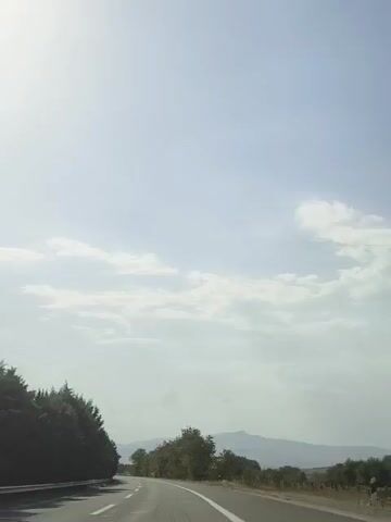 A time-lapse video shows a road curving to the right with trees on either side. The sky is bright and overcast with scattered clouds.
