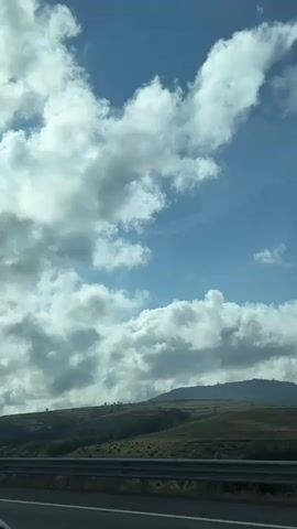 The video shows a view from a car traveling on a highway. The sky is blue with fluffy white clouds. In the distance, there are rolling hills covered in green vegetation. A guardrail runs along the side of the road. The car's rear end is visible in the lower left corner of the frame. The scene appears to be a sunny day with a light breeze.