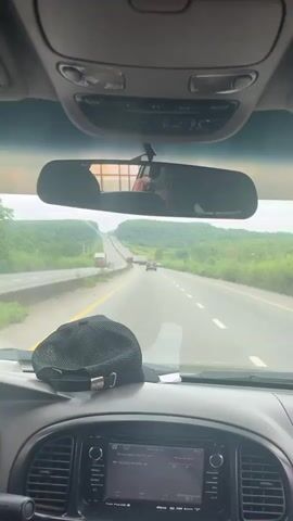A person records from the passenger seat of a car traveling on a multi-lane highway. The view is through the windshield, with the dashboard and rearview mirror visible. A black baseball cap rests on the dashboard. The highway stretches into the distance, with a truck and several cars ahead. Lush green vegetation lines both sides of the road. The sky is overcast.