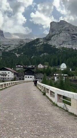 The video begins with a view of a cobblestone road leading uphill towards a village nestled in a valley. Mountains rise in the background, some with snow-capped peaks, and the sky is filled with dramatic clouds. The camera pans to the right, revealing a white railing along the road. As the video progresses, the perspective shifts to a wider view of the valley and the mountains, with a large body of water, likely a lake, visible in the foreground. The scene transitions to focus on the lake and the surrounding rocky slopes. The overall impression is of a serene mountain landscape with a man-made dam or bridge.