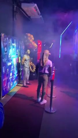 The video shows a crowded event venue, lit with blue and purple neon lights and filled with artificial fog. People are dressed casually and milling around, some taking pictures with their phones. There are stanchions with red ropes dividing areas, and a backdrop wall with logos is visible. Red and gold balloons add a celebratory touch. The floor appears to be dark and reflective. The music is loud and danceable.