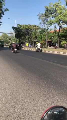 A street view video shows a road with vehicles and trees. In the foreground, a red side mirror reflects a person on a motorcycle. The road is asphalt and appears to be a street in a city or town. Several people on motorcycles are seen on the road, as well as a car. Trees line the side of the road, and some buildings are visible in the background. The weather appears to be sunny. The video is short, lasting only a few seconds.